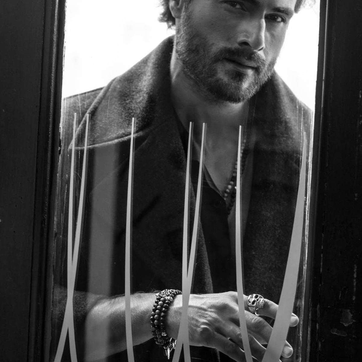 Black and white photo of a man wearing AN JEWELS JEWELRY Mod. AA.P255SBL bracelet and rings, looking through a stylized glass door.