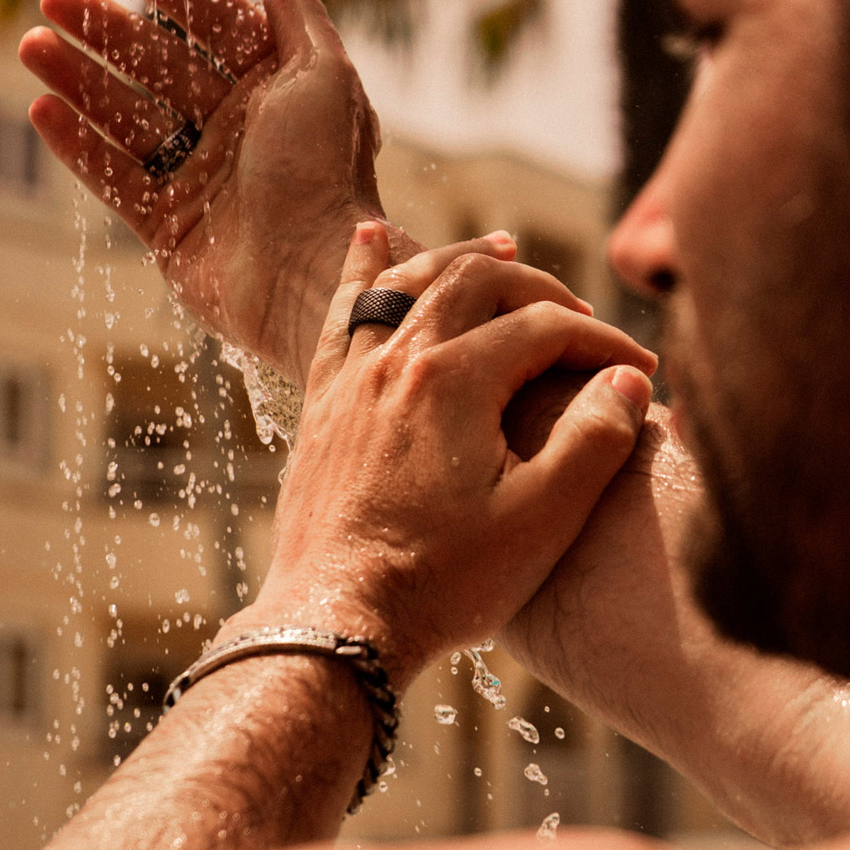 Man wearing rings and bracelet, washing hands under water spray in outdoor setting